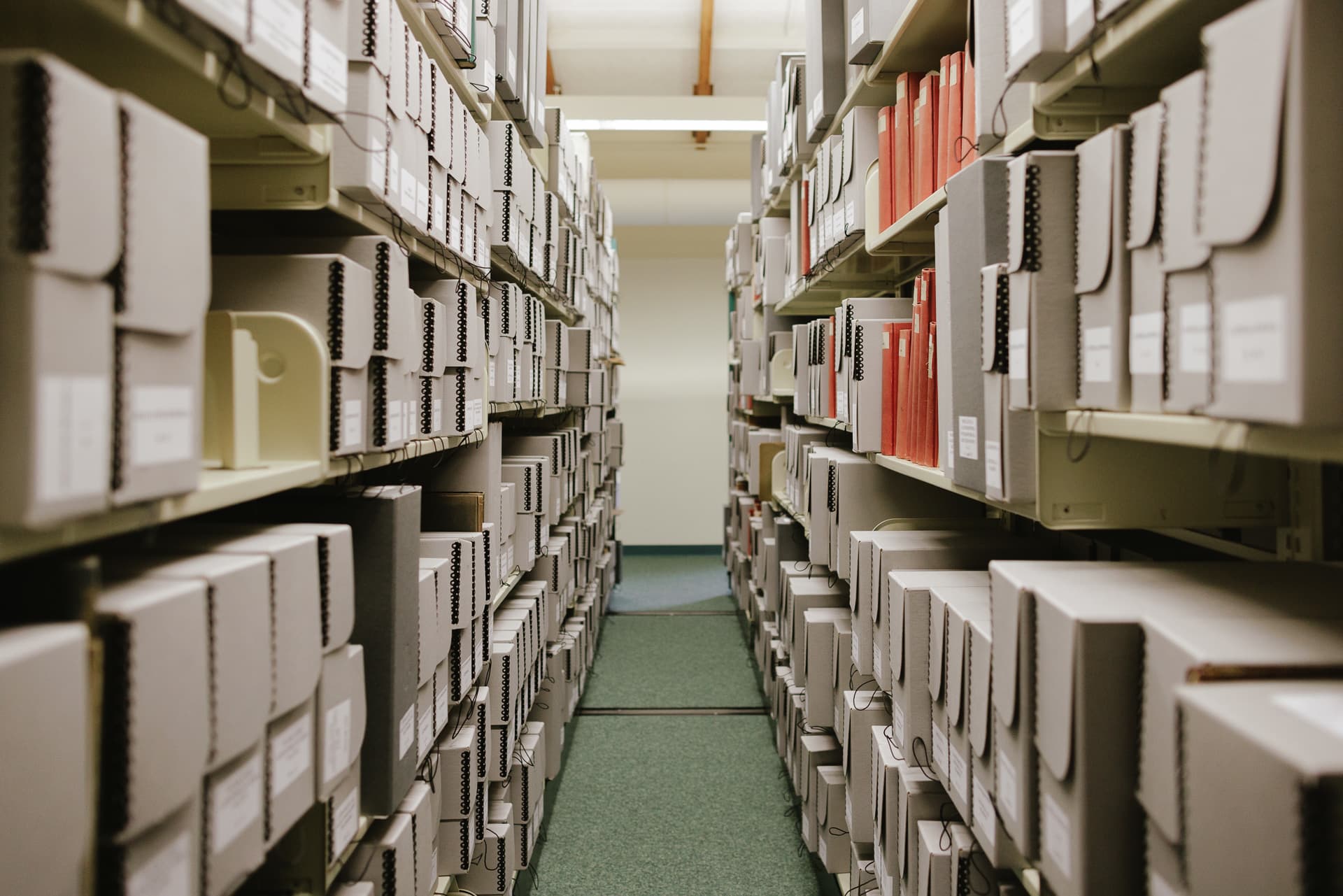 Looking down the aisle between two stacks of library shelving, the shelves are completely full of archival boxes and a few volumes of bound papers with red covers.